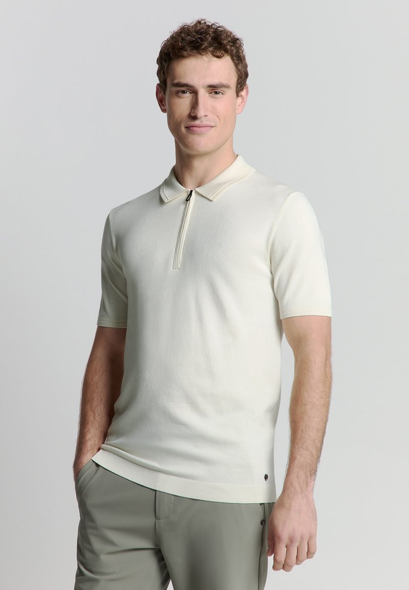 Korte mouwen polo shirt in crème-kleurige brei. Heeft een ritskraag en een subtiele ribbeltextuur. Gecombineerd met lichtgroene broek.