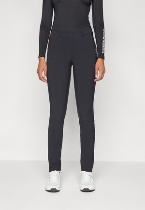Persona con pantalones ajustados negros, camiseta negra de manga larga y zapatillas blancas, de pie contra un fondo claro y liso.