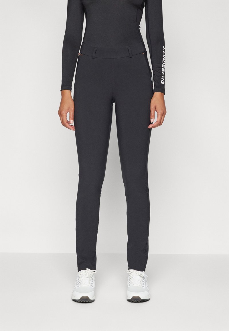 Personne portant un pantalon noir ajusté, un haut noir à manches longues et des baskets blanches, debout devant un fond clair uni.