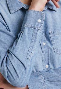 Personne portant une chemise boutonnée bleue claire à manches longues, bras plié et main reposant près du col.