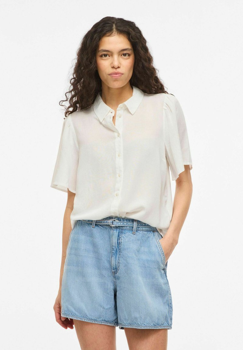 Femme aux cheveux bouclés portant un chemisier blanc à manches courtes rentré dans un short en jean bleu clair taille haute, une main dans la poche.