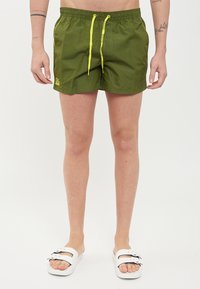 Shorts de bain verts avec une taille élastique, cordon de serrage jaune, poches latérales et un détail de logo. Portés avec des sandales blanches.