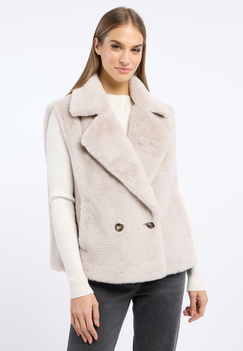 Kamizelka z futra sztucznego w jasnym beżu, dwurzędowy fason z dużym wykładanym kołnierzem i dwoma guzikami, w połączeniu z białym swetrem ryżowym.