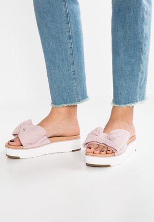 Voeten met lichtroze slip-on platformsandalen met strik, gecombineerd met cropped blauwe spijkerbroeken tegen een witte achtergrond.