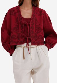 Rød cropped jakke med blomstermotiver, rund halsudskæring og båndlukning, parret med en ribbet top og cremede høj talje bukser.