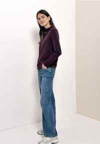 Dunkelrotes langärmeliges Oberteil kombiniert mit hellblauen gerade geschnittenen Jeans und silbernen Sportschuhen, stehend vor einem neutralfarbenen Hintergrund.