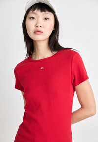 Camiseta corta roja de manga corta en tela de canalé con escote redondo y un pequeño detalle de logo plateado en el pecho. Diseño simple y ajustado.