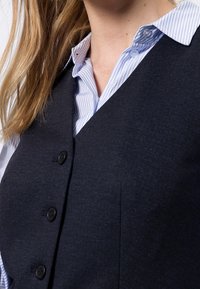 Ciemnoniebieska kamizelka z teksturowanej tkaniny, z czterema czarnymi guzikami, noszona na jasnoniebieskiej i białej koszuli w paski.