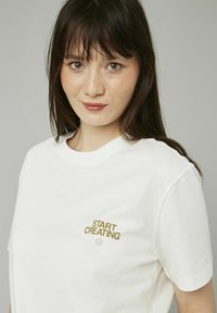 T-shirt en coton blanc à manches courtes, avec l'inscription "DÉBUTEZ LA CRÉATION" en doré et un graphique de fleur rose près du col.