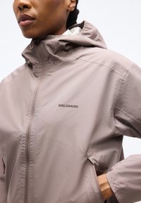 Personne portant une veste zippée taupe Salomon avec capuche, main dans la poche, légèrement tournée vers la gauche sur un fond uni.