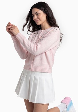 Giovane donna con i capelli lunghi e scuri che indossa un cardigan rosa, una gonna bianca plissettata e scarpe da ginnastica bianche con dettagli rosa e blu.