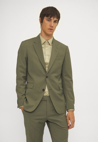 Olivgrüner Anzug mit passender Hose, einreihiger Jacke und TaschenDETAIL, kombiniert mit einem hellbeigen Hemd mit Kragen. Glatte Textur.