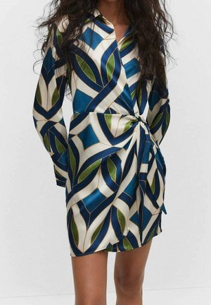 Frau trägt ein gemustertes Wickelkleid mit blau-grünen und weißen geometrischen Blättern, steht vor einem schlichten hellen Hintergrund.