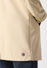 Μπεζ ελαφρύ μπουφάν με λείο υφή, πλευρικά κουμπιά και κυκλικό λογότυπο κοντά στο στρίφωμα. Κομψή εφαρμογή με μινιμαλιστικό σχέδιο.