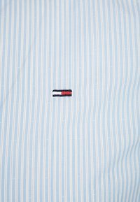 Ljusblå randig skjorttyg med subtil textur; har en liten broderad logotyp i rött, vitt och marinblått placerad centralt.