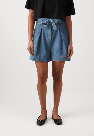 Femme portant un short en jean plissé taille haute avec une ceinture nouée, un haut noir et des ballerines noires, debout devant un fond uni.