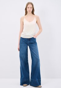 Beige v-neck camisole paired with wide-leg blue denim jeans, featuring pockets and a flared hem, displayed against a plain background.