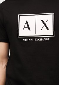 Μαύρο μπλουζάκι με λευκό ορθογώνιο λογότυπο που δείχνει "A | X" πάνω από το κείμενο "Armani Exchange" στο στήθος.