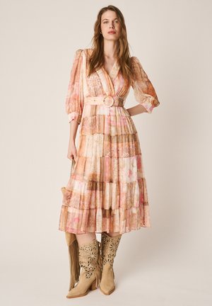 Donna in piedi che indossa un vestito midi con motivi, maniche a sbuffo, una cintura e stivali da cowboy beige con frange su uno sfondo neutro.