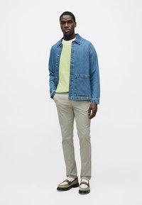 Giacca di jeans azzurro chiaro, indossata sopra un maglione verde testurizzato. Abbinata a pantaloni beige e scarpe multitone con suola spessa.