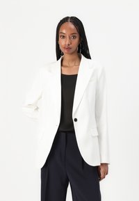 Bílý oblekový blazer s klopami a jedním černým knoflíkem. Nosený na černém topu, spárovaný s námořnickými kalhotami. Hladká textura.