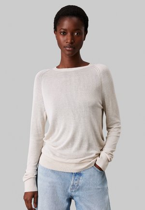 Maglione lungo beige chiaro a maniche lunghe con scollatura rotonda, polsini a coste e vestibilità rilassata, abbinato a jeans denim azzurri.