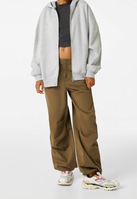 Persona vistiendo una sudadera gris con cremallera, un top negro corto, pantalones sueltos marrones y zapatillas blancas con acentos de colores, de pie sobre un fondo blanco.