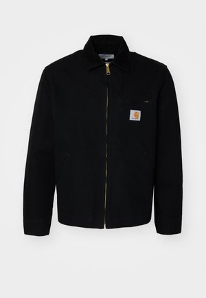Giacca nera in canvas con zip frontale, colletto a punta e tasche laterali. Presenta un piccolo logo sulla tasca del petto. Dettaglio della zip dorata.