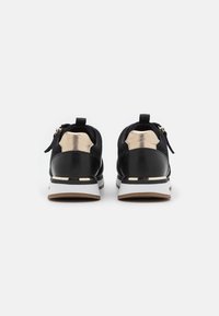 Un paio di sneakers nere con accenti dorati e suole bianche viste da dietro su uno sfondo bianco uniforme.