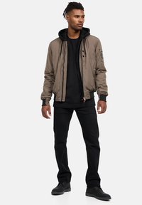 Giacca bomber marrone con zip frontale, polsini a coste e tasche laterali, indossata sopra un hoodie nero e pantaloni neri, abbinata a scarpe nere.