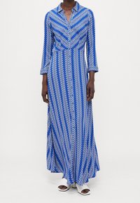 Długa niebiesko-biała sukienka w wzory z kołnierzykiem, z motywem chevronu i długimi rękawami, wykonana z lekkiego materiału.