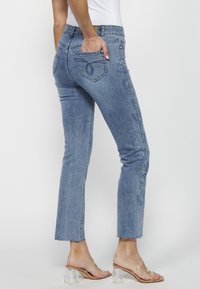 Personne portant un jean bleu clair brodé avec un ourlet brut et des talons à brides transparentes, main dans la poche arrière, debout devant un fond blanc.