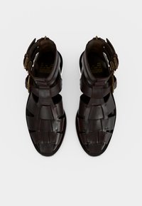 Σκούρα καφέ δερμάτινα σανδάλια με πλεκτούς ιμάντες και δύο χρυσά αγκράφες σε λευκό φόντο, από πάνω.