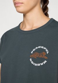 Dark green cotton t-shirt with a round neckline, featuring a brown tiger graphic and the text "PEACEFUL WARRIOR" in white circular design.