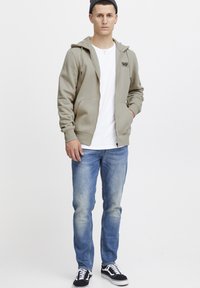 Sudadera beige con cremallera y bolsillos frontales, llevada sobre una camiseta blanca. Unos jeans de color azul claro y zapatillas negras completan el conjunto.