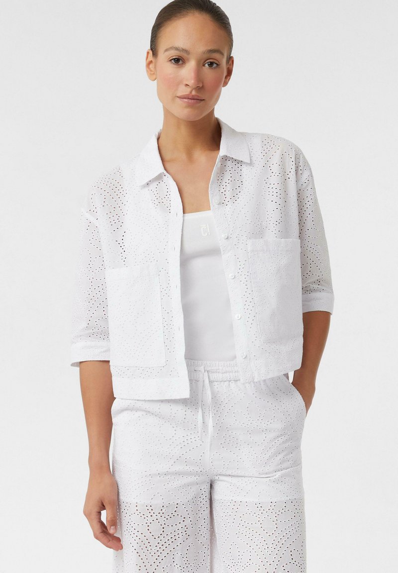Weiße Baumwollbluse mit Spitzen-Ausschnitt, Knopfleiste, zwei Vordertaschen und hochgekrempelten Ärmeln, kombiniert mit passenden weißen Spitzenhosen.