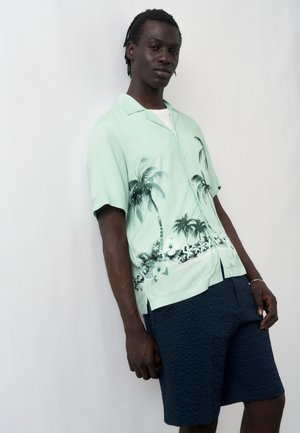 Man wearing light green short-sleeve shirt with palm tree print and navy textured shorts, standing against a plain white background.