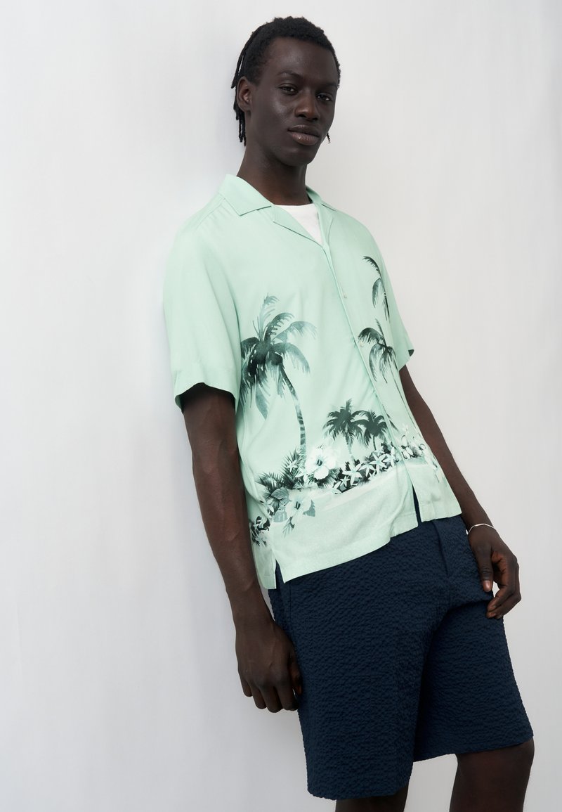 Άνδρας που φοράει ανοιχτοπράσινο κοντομάνικο πουκάμισο με σχέδιο από φοινικόδεντρα και σκούρο μπλε υφασμάτινες βερμούδες, στέκεται μπροστά σε απλό λευκό φόντο.