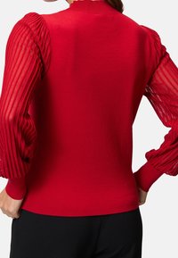 Czerwony sweter z gładkim dzianinowym korpusem i przezroczystymi, plisowanymi czerwonymi rękawami. Żebrowany kołnierz i mankiety dodają tekstury i detali do projektu.