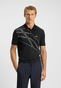 Czarna koszulka polo z białymi abstrakcyjnymi liniami. Krótkie rękawy, kołnierz i logo na piersi. Noszona z ciemnoniebieskimi spodniami i czarnym paskiem.