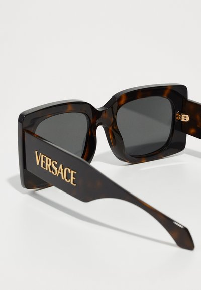 Čiliņš saulesbrilles ar kvadrātveida rāmjiem un tumšiem stikliem; sānos ir zelta "VERSACE" logotips, ar gludu spīdīgu apdari.