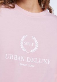 Růžové bavlněné tričko se bílým potiskem a designem vavřínu s nápisem "SET URBAN DELUXE SINCE 2009." Hladký materiál a výstřih do kulata.