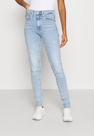 Jeans ajustados de color azul claro, fabricados en denim, con cintura alta, un ligero desgaste y diseño de cinco bolsillos, combinados con zapatillas blancas.