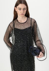 Rochie neagră cu paiete, cu mâneci lungi transparente, având un gât rotund. Asortată cu o mică geantă plic neagră și bleumarin.