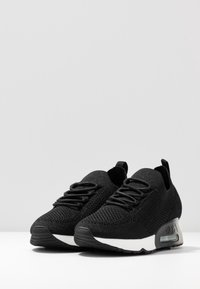 Paire de baskets en tissu tricoté noir avec des lacets noirs et des semelles blanches, présentées sur une surface blanche devant un fond gris clair.