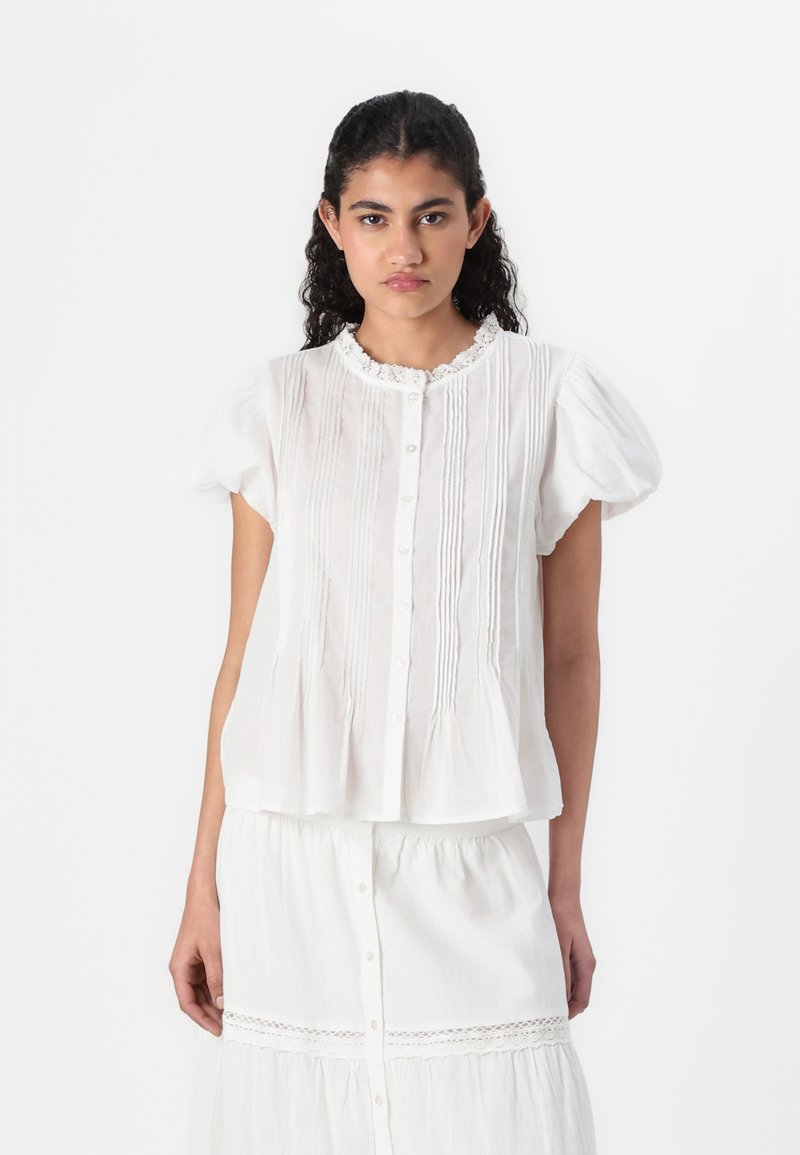 Hvit kortermet bluse med knapper foran, blondekrage, plissédetaljer og flared søm. Stoffet ser lett og teksturert ut.
