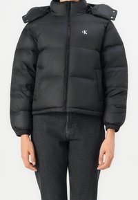 Veste matelassée courte noire avec fermeture éclair devant et petit logo blanc CK sur la poitrine, portée avec un jean taille haute gris foncé.