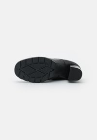 Zapato de cuero negro tumbado de lado, mostrando la suela texturada con un patrón de tracción y un tacón bajo apilado sobre un fondo blanco.