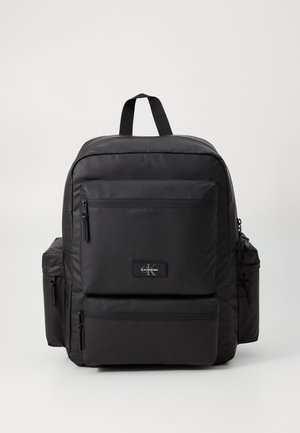 Sac à dos en nylon noir avec plusieurs poches zippées, bretelles rembourrées et une poignée supérieure. Il présente un patch logo à l'avant. Design simple.