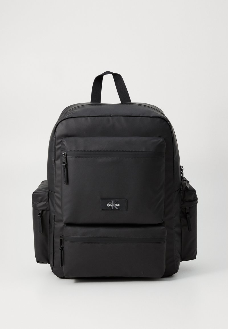 Black nylon backpack with multiple zippered pockets, padded straps, and a top handle. Features a logo patch on the front. Simple design.